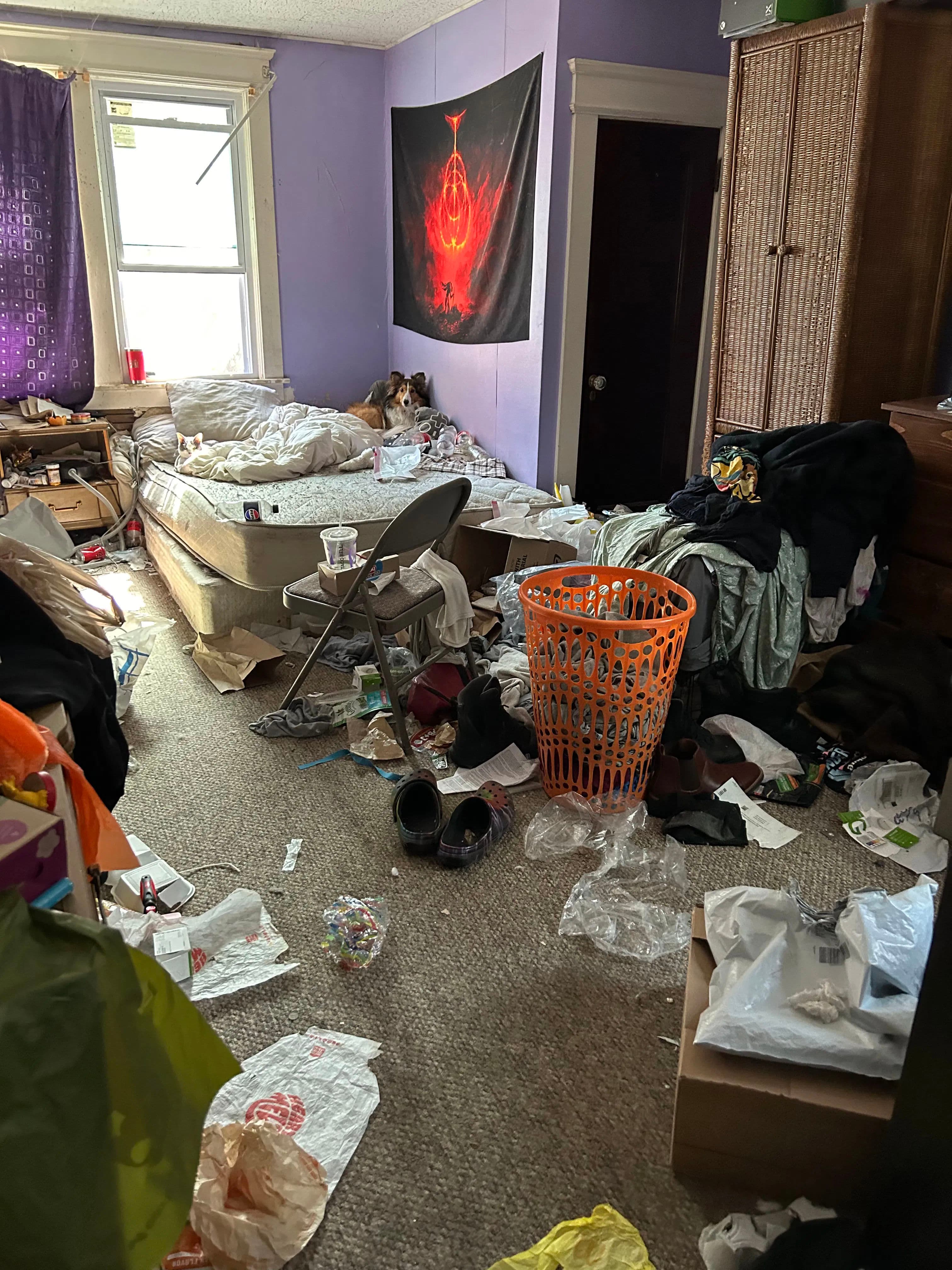Cluttered bedroom with messy bed, clothes, trash, and an orange laundry basket.